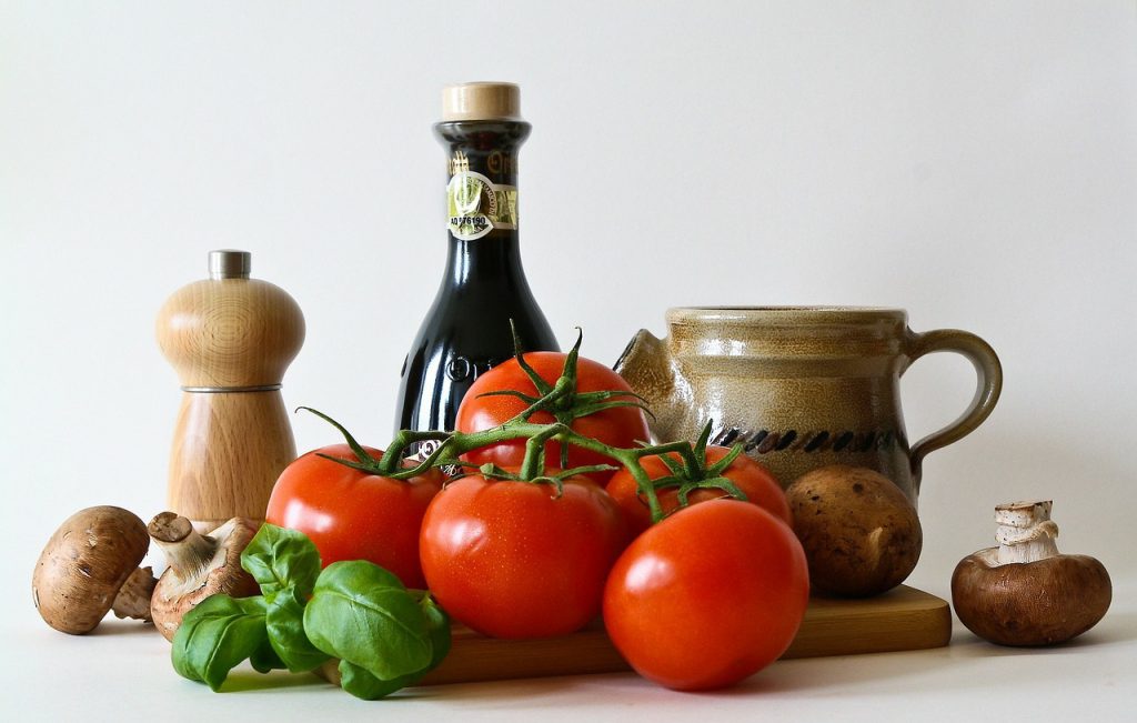 Kuvassa vihannes- ja ruoka-asetelma. Tomaattia, basilikaa, sieniä, pippurimylly ja öljypullo leikkuulaudalla