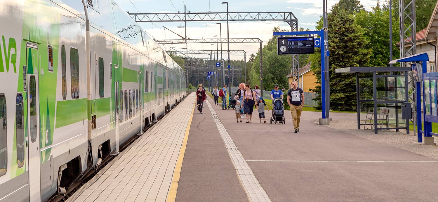 Juna Kannuksen rautatieasemalla, matkustajia kävelee laiturilla, opastauluja ja aseman rakennuksia.