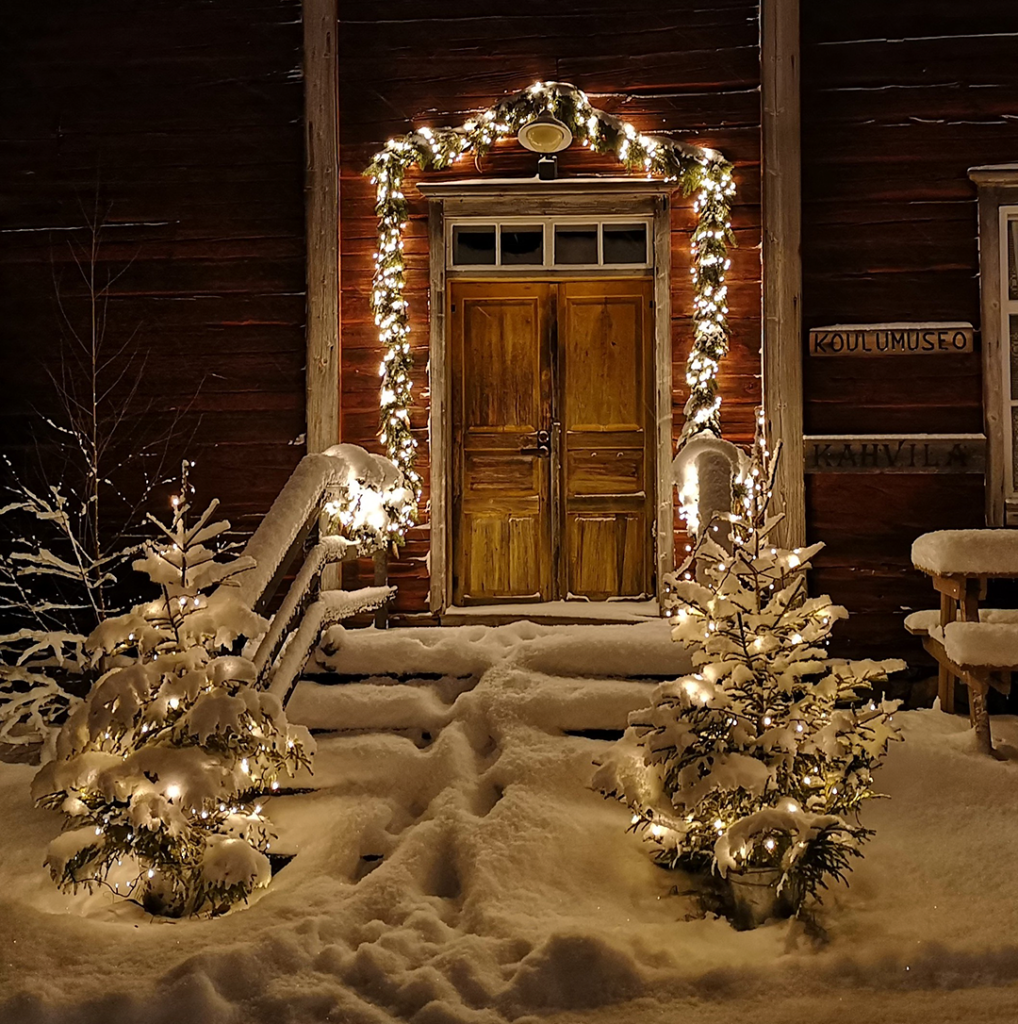 Jouluvaloin ympäröity vanhan punaisen hirsirakennuksen sisäänkäynti, luminen maisema, pimeä ilta
