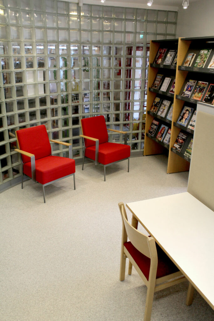 Kaksi punaista nojatuolia, lasitiiliseinä, lehtilyjä ja lukupöytä.